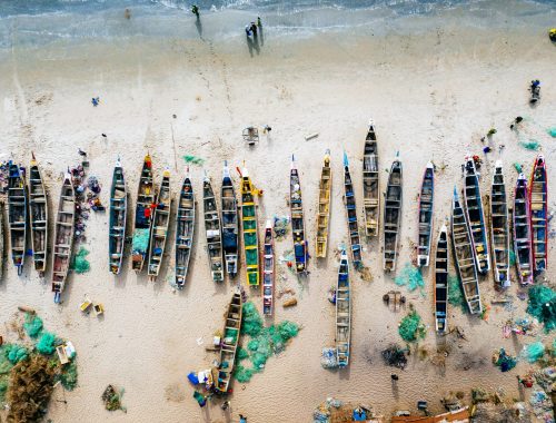 An overhead aerial shot of different colored boats on a sandy beach with the sea nearby